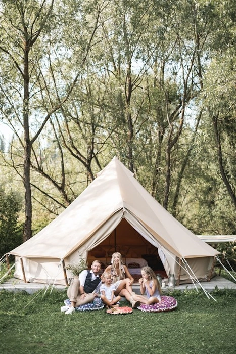 A family camping with children sitting on the grass in front of a large bell tent surrounded by trees