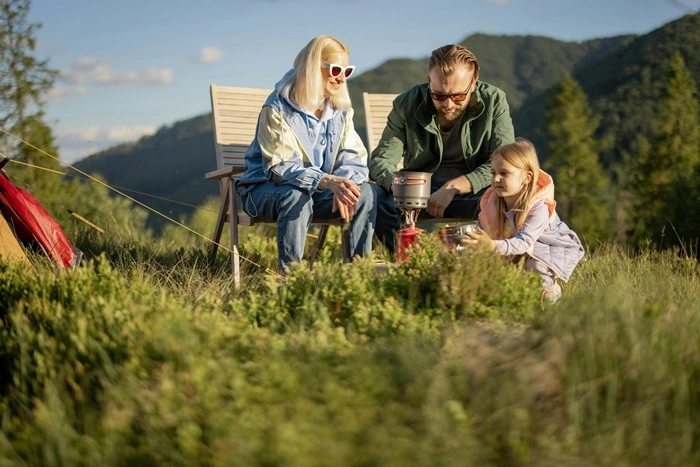 A family with a young girl cooking outdoors on a camping stove with a tent and mountain scenery behind them