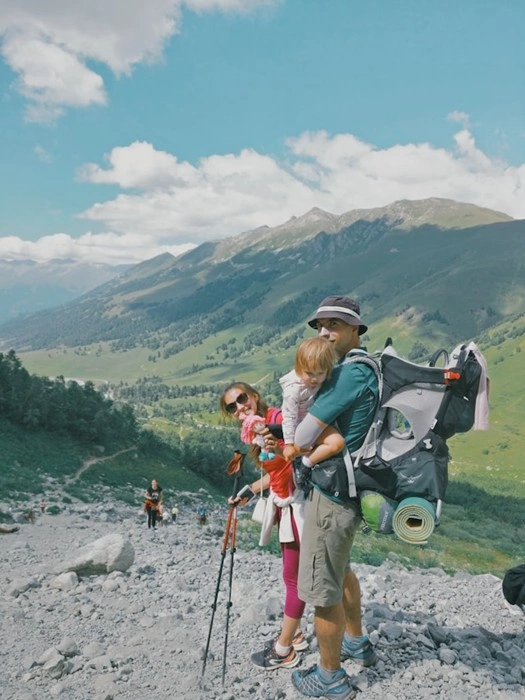 Parents hiking with a young child in a backpack carrier on a mountain trail with stunning alpine scenery
