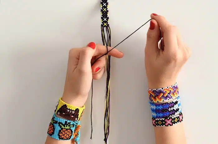 Two hands weaving a friendship bracelet with colorful woven bracelets on both wrists, representing the bracelet scam that targets tourists in popular destinations.