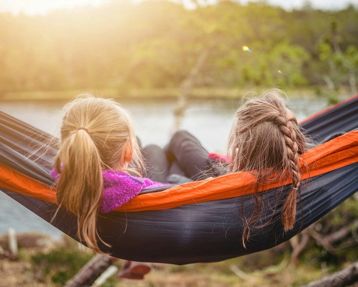 Two young girls lying in an orange and black hammock overlooking a calm lake with trees and warm sunset light
