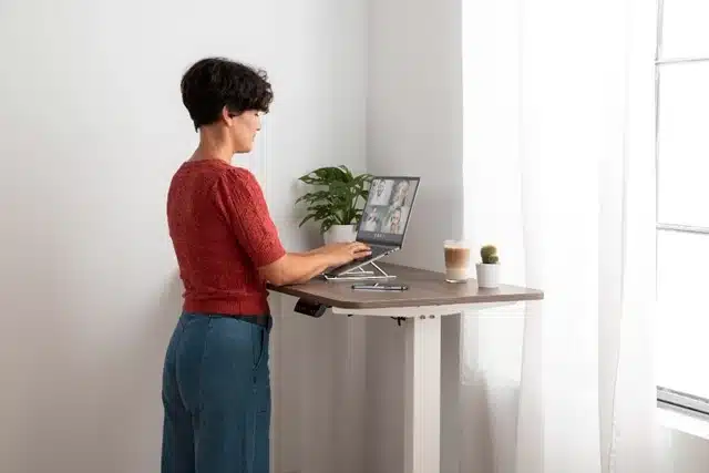 Student on video call using laptop stand at standing desk with plants and coffee nearby
