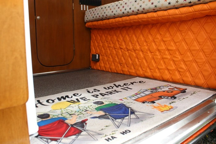 "Home is where we park it" doormat inside a cozy RV with orange quilted seating and a fold-out bed
