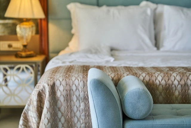 A neatly dressed hotel bed with a textured gold duvet, layered white pillows, and a decorative bolster cushion in the foreground