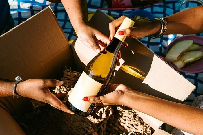 Two people carefully placing a white wine bottle into a cardboard box with protective shredded paper padding