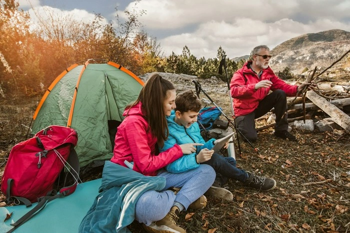 A mother and young boy looking at a tablet together outside a green tent while an older man gathers firewood in a mountain setting