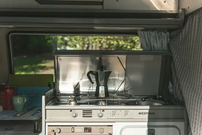 why motorhome travel works — a compact Westfalia kitchen with a moka coffee pot, gas stove, and small sink surrounded by greenery