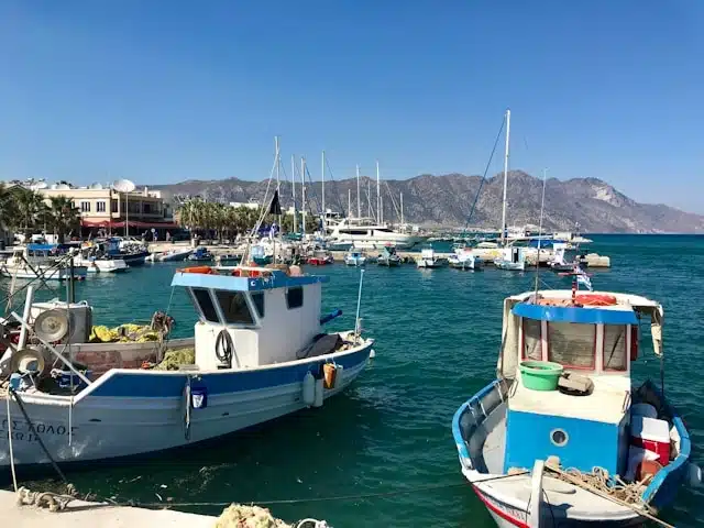 Traditional fishing boats and sailboats anchored in a small harbor in Kos, Greece, with turquoise water and mountain backdrop