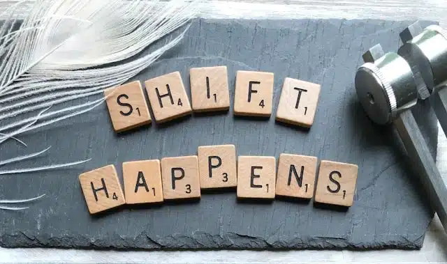 Scrabble tiles spelling out "Shift Happens" on a dark slate surface next to a feather and a metal object