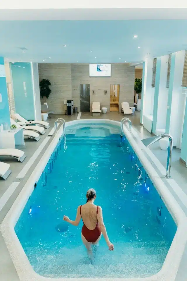 Woman wading into an indoor spa pool at an upscale wellness clinic with loungers and steam rooms