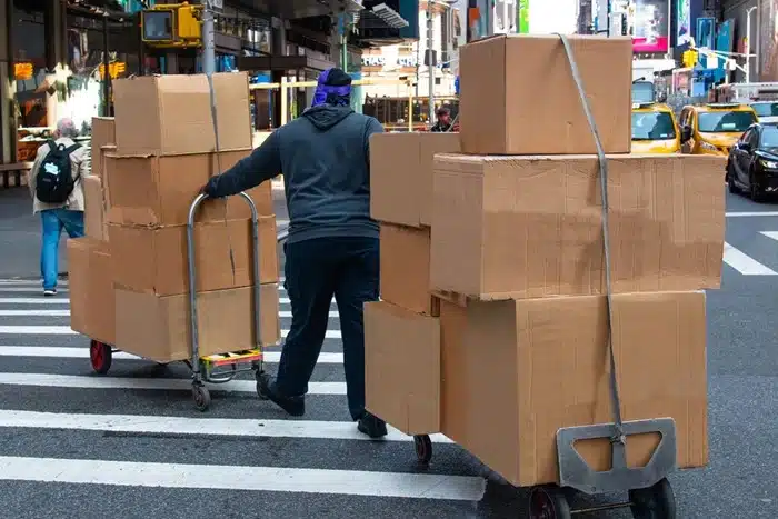 Man pushing a hand truck stacked with cardboard boxes through a busy NYC street
