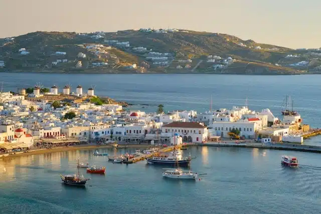 Aerial view of a whitewashed Greek island village at golden hour with sailboats dotting the calm harbor waters