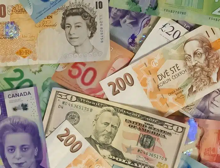 A spread of various international banknotes including US dollars, British pounds, Canadian dollars, and Czech koruna, representing the risk of money exchange scams in tourist areas.
