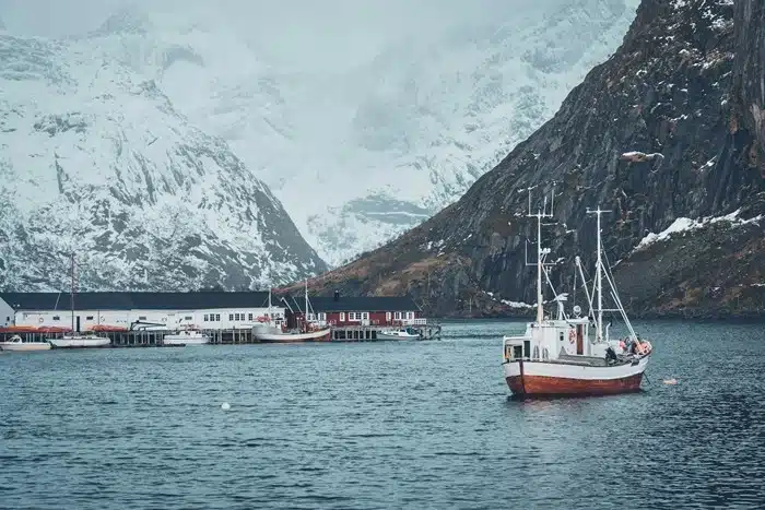 a fishing boat anchored in a Norwegian fjord village with snow-capped mountains and red waterfront buildings in the background