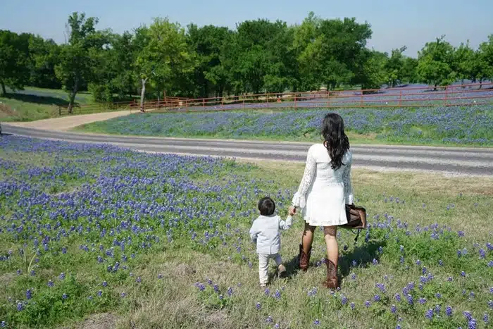 A woman in a white dress and cowboy boots holding hands with a toddler while walking through a field of bluebonnet wildflowers on a Texas Hill Country getaway