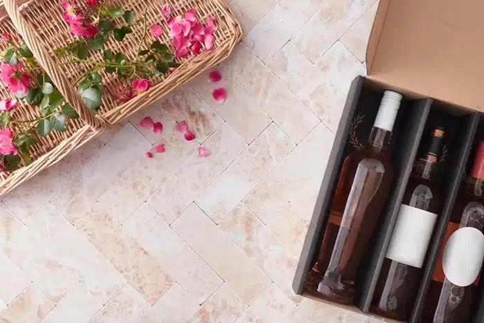 Multiple wine bottles secured in a padded gift box on a tiled surface next to a wicker basket with pink flowers