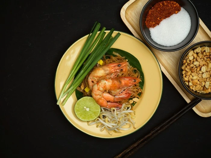 Overhead view of a traditional Thai pad thai with prawns, rice noodles, lime, and condiments on a dark background