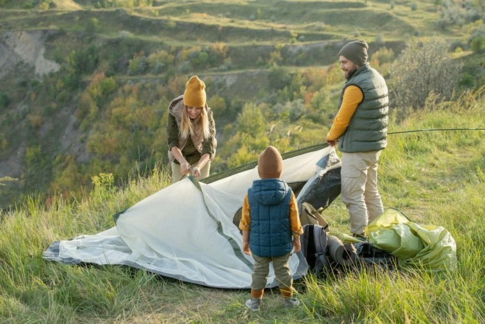 Parents and a small child pitching a tent on a green slope with rolling hills and trees in the background