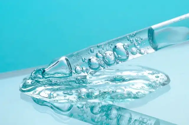 A dropper dispensing clear peptide serum with bubbles on a reflective surface against a blue background