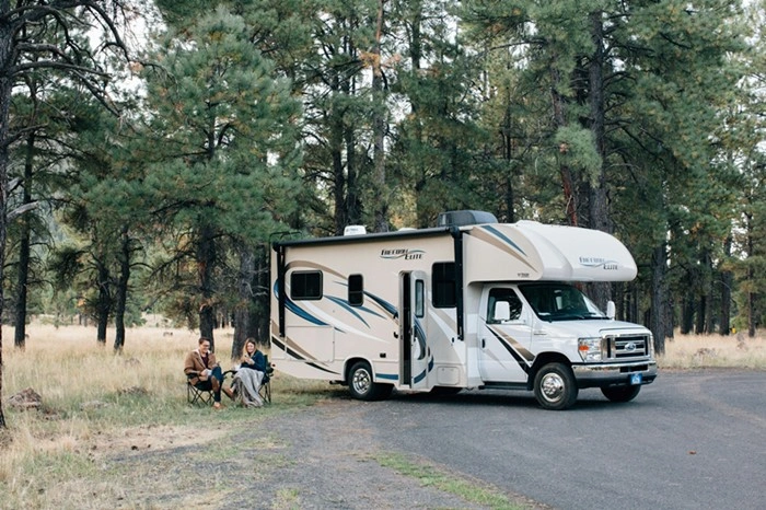 Couple relaxing in camping chairs beside a large RV parked among pine trees in a quiet forest campground