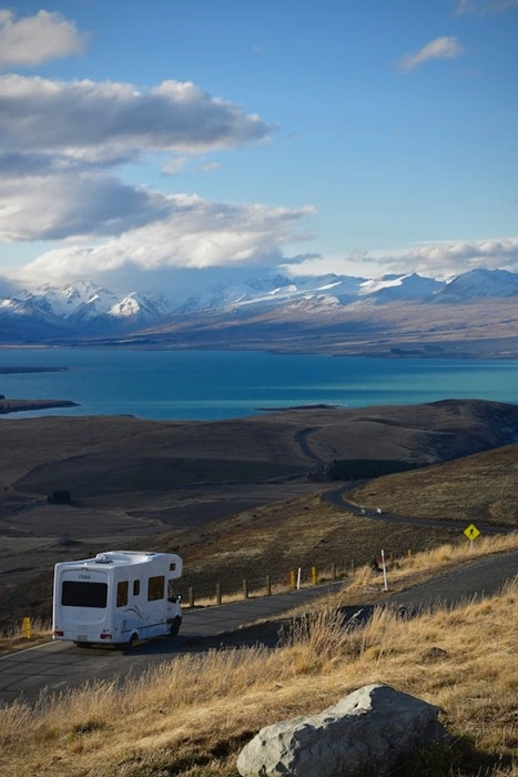 White RV driving along a scenic mountain road with a vast turquoise lake and snow-capped peaks in the distance