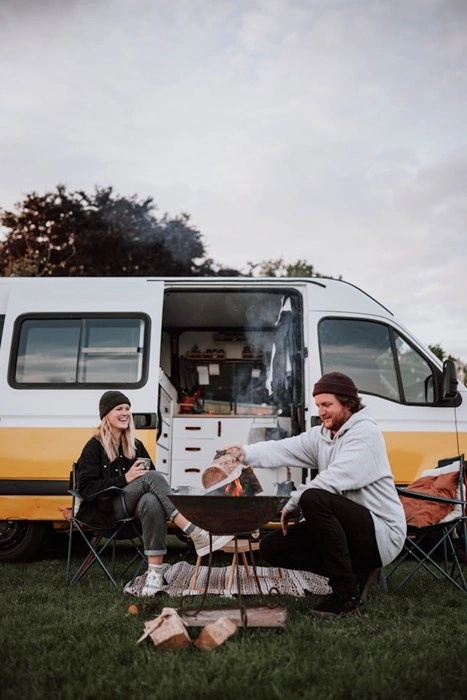 Couple laughing and tending a campfire in front of their converted camper van parked on a grassy site