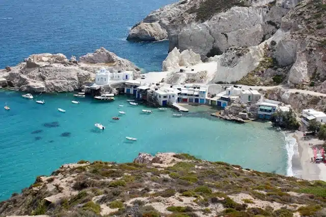 Aerial view of a secluded Greek coastal village tucked into white volcanic cliffs, with small boats floating in vivid turquoise water