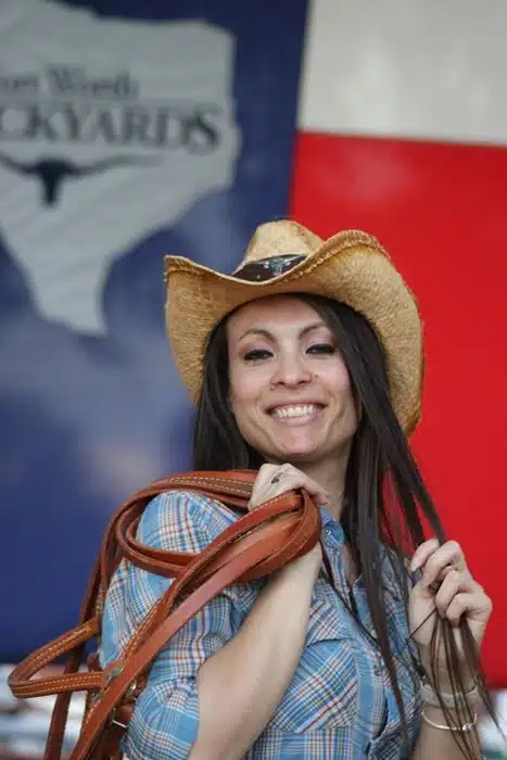 A smiling woman in a straw cowboy hat and plaid western shirt holding leather reins in front of a Texas flag at a Fort Worth Stockyards event