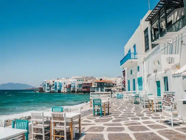 Waterfront cafe tables and chairs lining the cobblestone promenade of Little Venice in Mykonos, with whitewashed buildings meeting the Aegean Sea