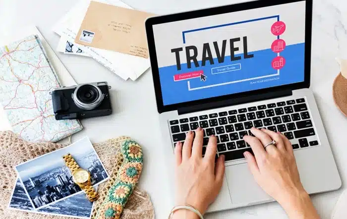 A traveler browsing a travel website on a laptop with a camera, printed photos, and maps nearby, illustrating the risk of fraudulent travel booking and review platforms.