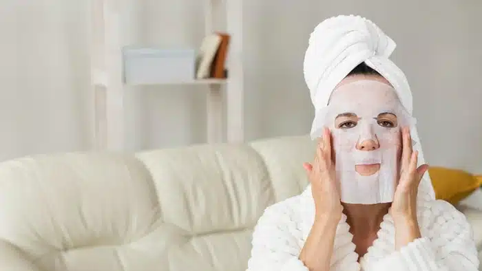 a traveler in a bathrobe applying a sheet facial mask for skin hydration