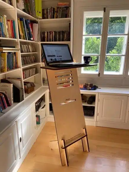 Slim wooden standing desk with laptop set up beside a window in a home library