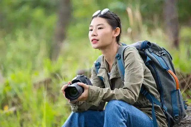 A woman hiker with a camera and backpack resting outdoors in a lush green forest during an adventure trip