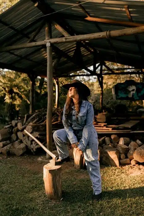A woman in a denim jacket and wide-brim black cowboy hat sitting on a log stump near a rustic woodpile at golden hour