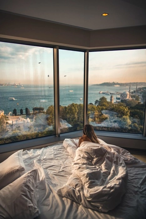 A woman sitting up in a white hotel bed surrounded by soft rumpled bedding, gazing out at a stunning sea and city view through floor-to-ceiling corner windows