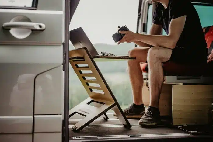 man working on a laptop with a camera in hand inside a motorhome with scenic countryside views outside