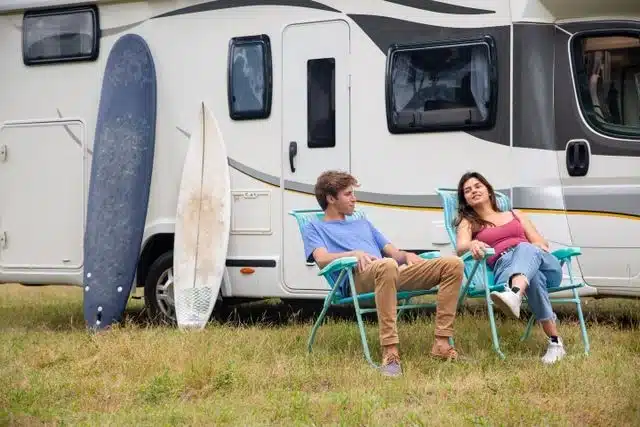 Couple relaxing in camping chairs beside their luxury camping trailer with surfboards
