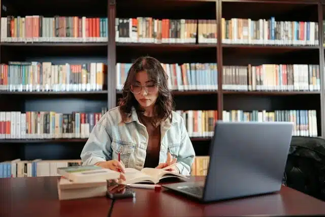 Student studying with laptop and books in library, pursuing online engineering degree