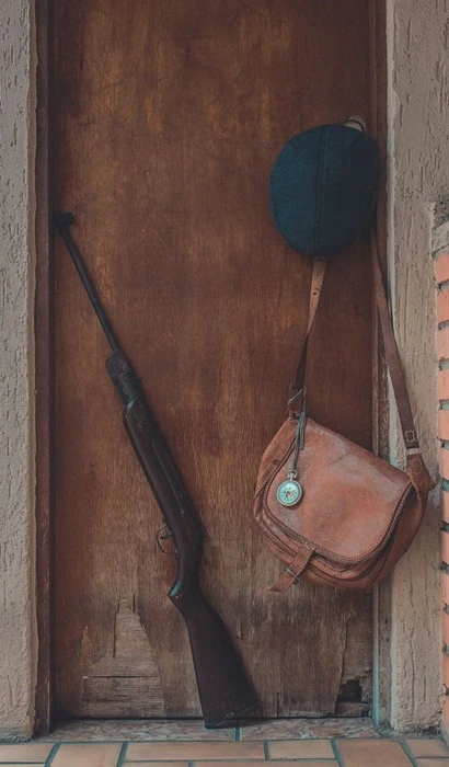 A rifle leaning against a wooden door beside a leather travel bag and hat, evoking concealed carry gear ready for a road trip