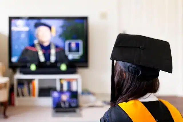 Graduate wearing cap watching virtual degree ceremony on TV screen