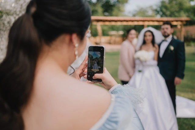 Wedding guest photographing bride and groom outdoors, capturing candid event photo moments