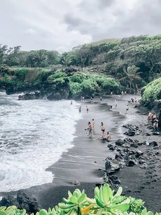 Visitors relaxing on a black sand beach surrounded by lush greenery in Maui