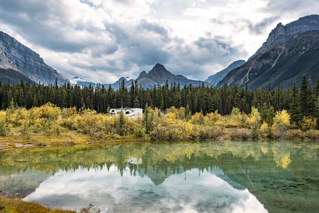 RV parked near a mirror-like lake with mountain peaks and autumn trees reflected in the water, Alberta Canada