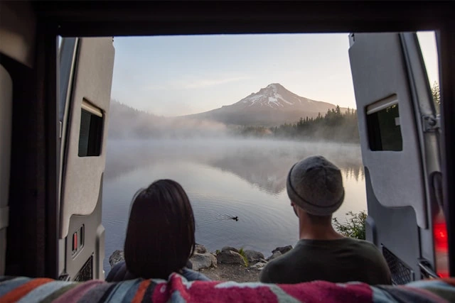 Couple enjoying morning travel views of a misty mountain lake from the open back doors of their RV
