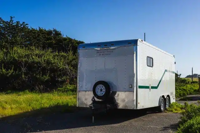 Enclosed camping trailer with a mounted spare tire, parked in a scenic outdoor spot