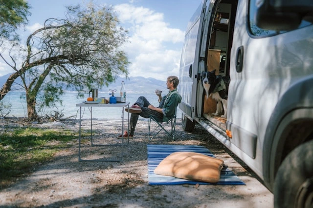 Traveler relaxing in a camp chair by the coast with his dog watching from inside the RV van