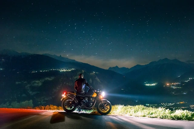 Rider pausing on a mountain road at night, motorbike headlights illuminating a starlit valley view
