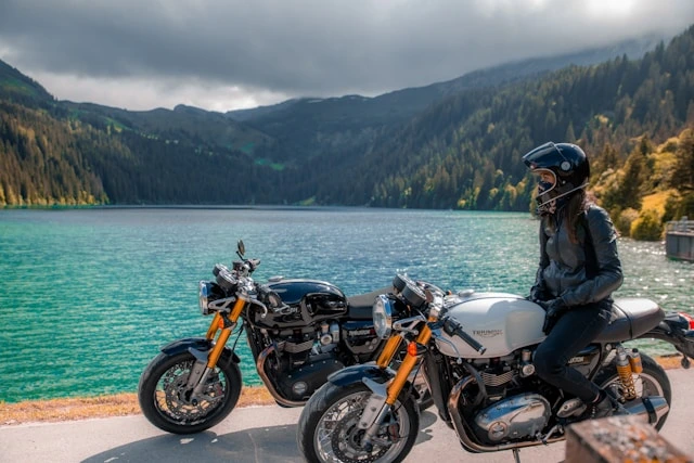 Rider seated on a motorbike beside a turquoise mountain lake with two more bikes parked nearby