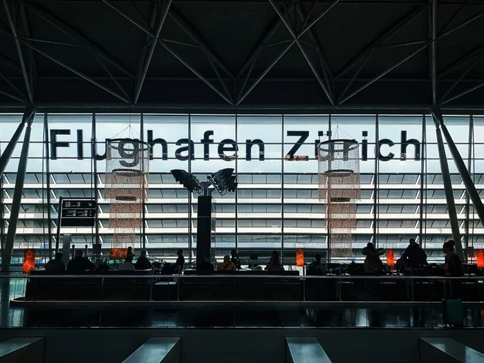 Passengers waiting in the boarding lounge at Zurich Airport (Flughafen Zürich), with travelers seated by the departure boards.
