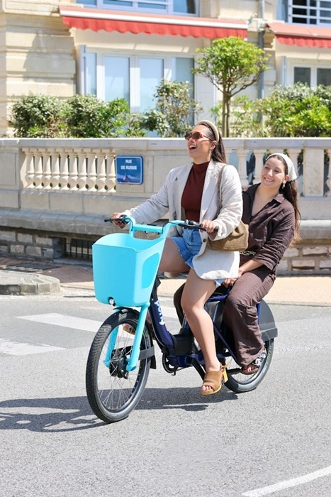 Two women riding an e-bike together, one seated on the rear rack, laughing on a sunny street